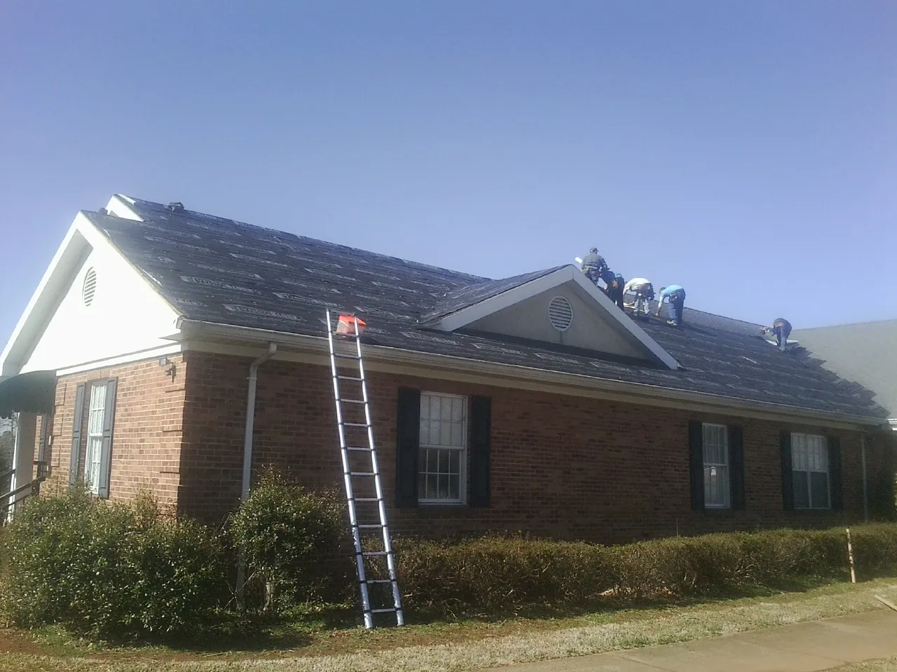 Crew tearing shingles off a brick ranch roof with a ladder set — Emergency Roof Repair work by our crew in Horicon