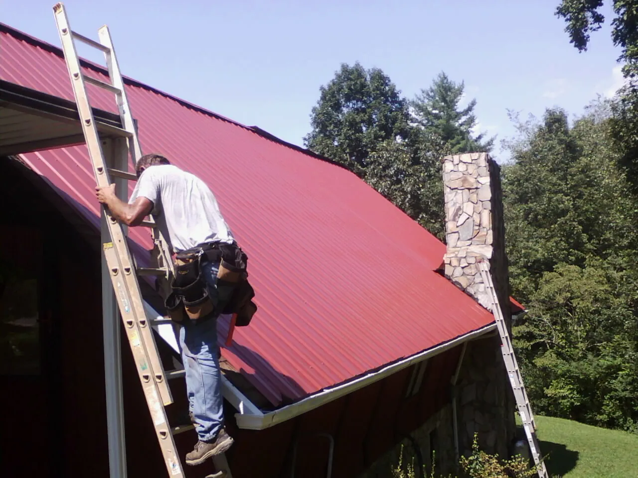 Roofer on a ladder beside a red metal roof and stone chimney — Soffit & Fascia Repair by Roofing Brothers in Horicon